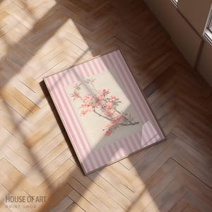 May include: A framed floral print featuring a branch with pink blossoms. The artwork is set against a cream background with a pink striped border. The print is displayed on a wooden floor, with the text "HOUSE OF ART PRINT SHOP" visible.