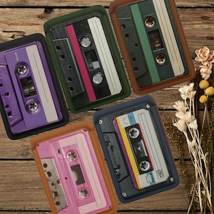 Può includere: Una vista dall'alto di cinque musicassette in vari colori, tra cui viola, rosa, verde e blu, ciascuna in una custodia protettiva. Le musicassette sono disposte su una superficie di legno, con fiori secchi sullo sfondo.