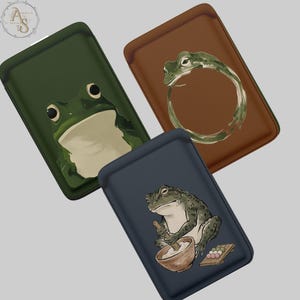 Puede incluir: Tres fundas de cuero para teléfono con ilustraciones de ranas. Una es verde oscuro con una cara de rana, otra es marrón con una rana en un círculo, y la tercera es azul marino con una rana preparando comida. Cada funda tiene un borde cosido.