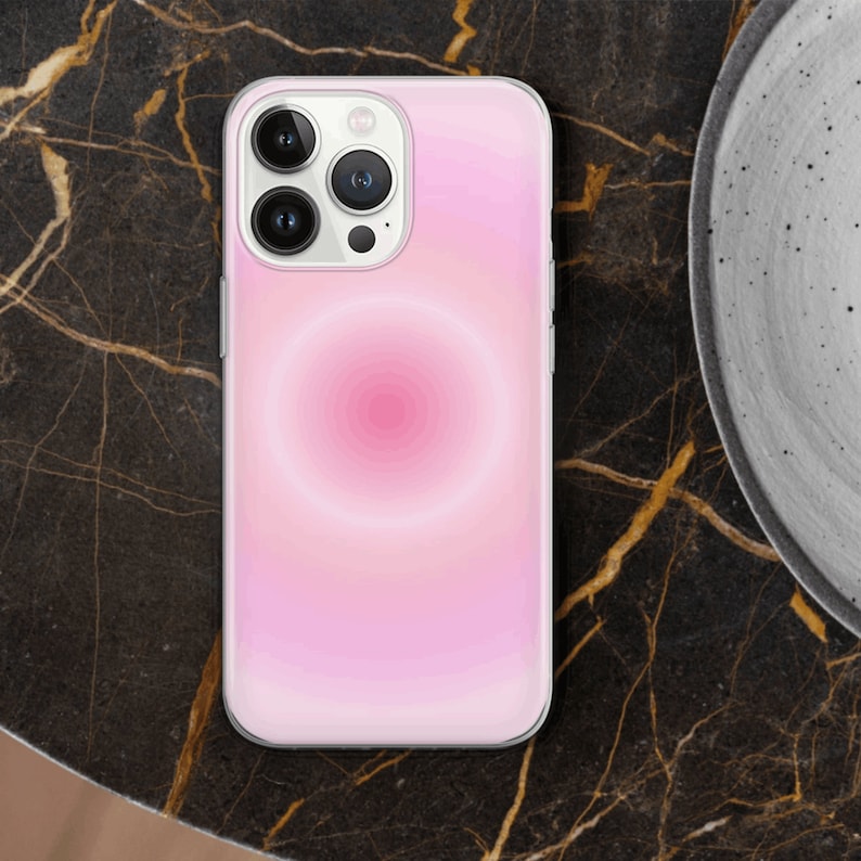 May include: A white iPhone case with a pink and white gradient design. The case has a circular pattern in the center and a camera lens cutout.