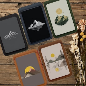 以下が含まれることがあります： 風景画が描かれた長方形のケースが5つ。ケースは革のような外観で、さまざまな色の縁があります。アートには山脈、太陽、月が含まれています。1つのケースには「Take a breath」というテキストがあります。