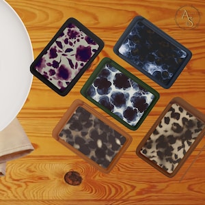 May include: A collection of rectangular trays with various patterns, including floral, abstract, and animal prints. The trays have black, green, and brown frames, and are arranged on a wooden surface.