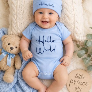 May include: A light blue baby bodysuit and matching beanie set. The bodysuit features the text "Hello World" embroidered in dark blue script on the chest. The beanie also features the text "Hello World" embroidered in dark blue script.