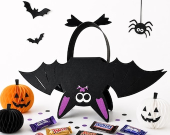 Może przedstawiać: Czarna torba na słodycze na Halloween w kształcie nietoperza z fioletowymi akcentami, otoczona papierowymi nietoperzami, dyniami, pająkiem, batonami czekoladowymi i lizakami. Torba ma uchwyt i jest ozdobiona twarzą.