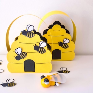 May include: Yellow paper beehive-shaped boxes with black and white bee cutouts. The boxes have a black entrance and are decorated with yellow and black paper bees. A yellow toy bee with a clear wing is also present.