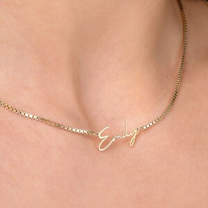 May include: A gold-colored necklace with the name "Emily" in a delicate script. The necklace is worn around the neck, showcasing the personalized pendant. The chain has a box chain design.