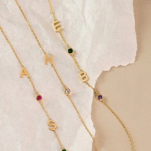 Op de afbeelding: Drie gouden kettingarmbanden met letterbedels en gekleurde edelstenen. De armbanden bevatten de letters A, S, B, E en N met rode, groene en paarse edelstenen.