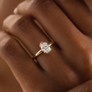 May include: A gold ring featuring a single oval-cut clear gemstone set in a partial bezel setting. The band is smooth and polished, showcasing a clean, minimalist design that highlights the central stone. The metal has a warm, yellow gold tone.