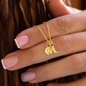 May include: A gold necklace featuring two charms: a circular pendant and a small wing. The necklace is suspended from a delicate chain. The jewellery is held up by a person's hand.