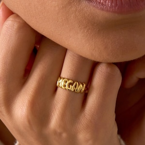 May include: A gold-colored ring with the name "MEGAN" spelled out in capital letters. The ring is worn on a finger. The ring is a simple band with the letters connected. The background is blurred.