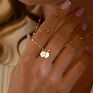 Könnte beinhalten: Eine zarte goldene Halskette mit zwei kleinen, runden Anhängern, die mit den Buchstaben "K" und "E" graviert sind. Die Halskette hat eine feine Kette mit kleinen, gleichmäßig verteilten Perlen. Der Schmuck wird auf der Haut einer Person präsentiert.