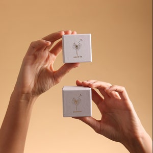 Puede incluir: Dos cajas cuadradas blancas con un dise&ntilde;o de diente de le&oacute;n y la palabra "GOLDMIN" impresa en ellas. Las cajas est&aacute;n siendo sostenidas por dos manos.