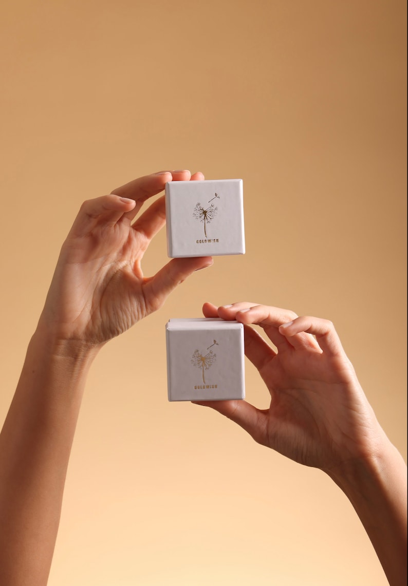 Puede incluir: Dos cajas cuadradas blancas con un dise&ntilde;o de diente de le&oacute;n y la palabra "GOLDMIN" impresa en ellas. Las cajas est&aacute;n siendo sostenidas por dos manos.