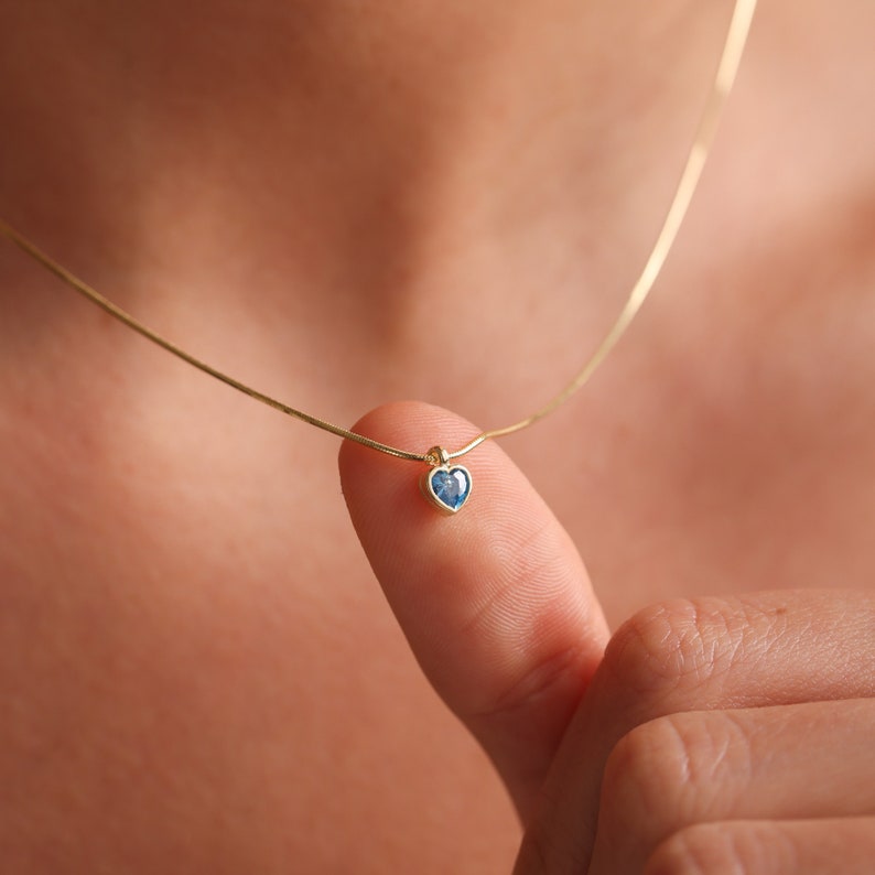 Puede incluir: Un delicado collar de cadena de oro con un peque&ntilde;o colgante de coraz&oacute;n con una piedra preciosa azul.