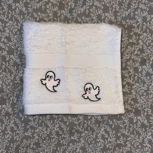 May include: White hand towel featuring two embroidered ghost designs. The ghosts have black outlines and red mouths. The towel is folded and has a textured finish. The background is a grey floral pattern.