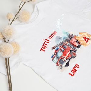 Könnte beinhalten: Weißes T-Shirt mit einem roten Feuerwehrauto und dem Text "Tatu Tata!" und "Lars" darauf gedruckt. Das Feuerwehrauto hat eine Leiter und einen Schlauch. Neben dem Feuerwehrauto befindet sich eine Nummer 4 in orangefarbenen Flammen.