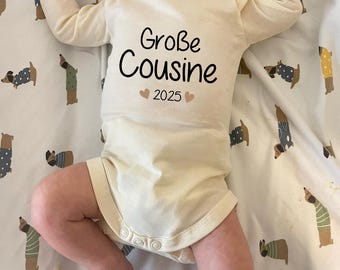 May include: A white long-sleeved onesie with the text "Große Cousine 2025" in black with two small pink hearts on either side of the year.
