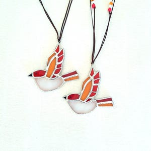 Peut inclure: Deux colliers en verre coloré en forme d'oiseau avec des accents rouges, orange et blancs. Les colliers sont sur des cordons bruns avec des perles orange et blanches.