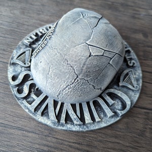 Może przedstawiać: Srebrny podkładka pod piwo Guinness z teksturowanym, popękanym wzorem. Podkładka zawiera logo Guinness i napisy "Dublin" oraz "St. James's Gate Brewery". Projekt zawiera symbol harfy.