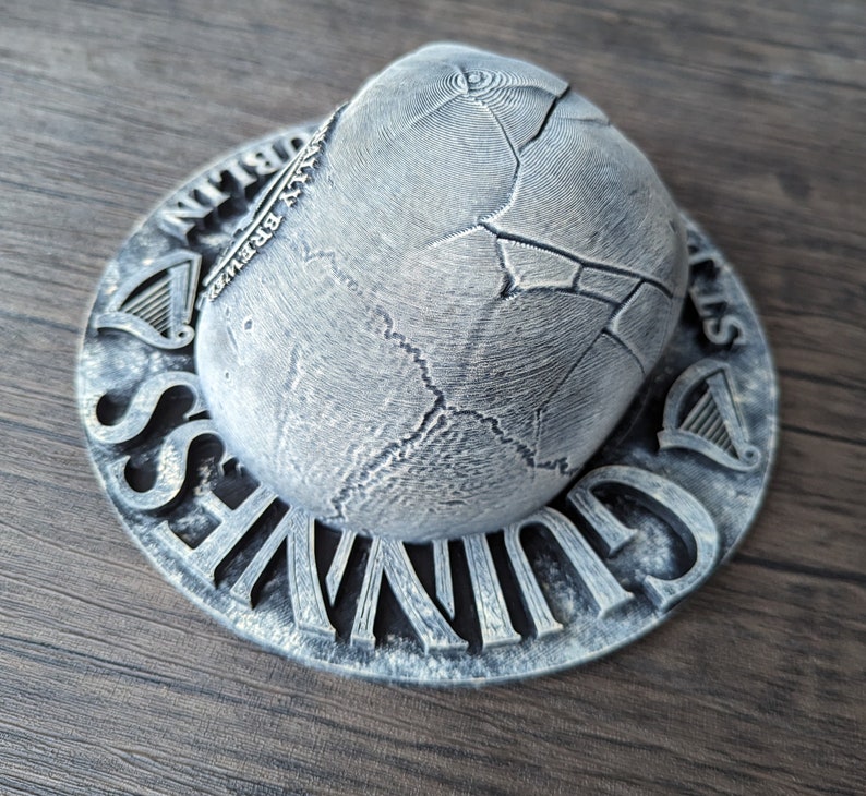Może przedstawiać: Srebrny podkładka pod piwo Guinness z teksturowanym, popękanym wzorem. Podkładka zawiera logo Guinness i napisy "Dublin" oraz "St. James's Gate Brewery". Projekt zawiera symbol harfy.