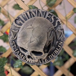 Może przedstawiać: Okrągła, wyglądająca na zwietrzałą tablica z motywem czaszki i logo Guinness. Tablica jest w odcieniach szarości i brązu, z napisami "Guinness", "St. James's Gate Dublin" i "Traditionally Brewed".