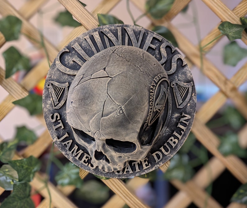 Może przedstawiać: Okrągła, wyglądająca na zwietrzałą tablica z motywem czaszki i logo Guinness. Tablica jest w odcieniach szarości i brązu, z napisami "Guinness", "St. James's Gate Dublin" i "Traditionally Brewed".
