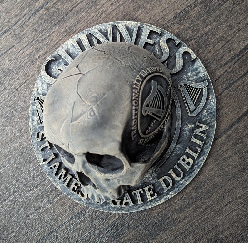Może przedstawiać: Szara, dekoracyjna czaszka z logo Guinness i tekstem. Okrągły wz&oacute;r zawiera słowa "Guinness", "St. James's Gate Dublin" i "Traditionally Brewed". Czaszka ma pęknięcia i szczeg&oacute;łowe cechy.