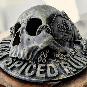 以下が含まれることがあります： 「The Kraken Black Spiced Rum」と書かれたラベルが付いた、3Dプリントされた頭蓋骨と触手。頭蓋骨は濃いグレーで、触手は薄いグレー。ラベルは黒地に白文字です。