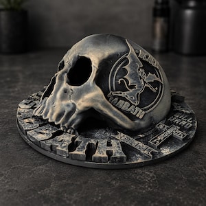 Puede incluir: Una escultura de calavera gris metálico desgastado con las palabras "Black Sabbath" y "Death" inscritas en la base. La base también presenta una cruz y el nombre "Bill Ward".