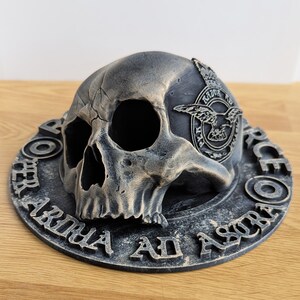 May include: A black and gold skull with a raised inscription that reads "FOR THE ARM OF ASRA" and a small emblem with a bird and the text "AIR FORCE" on the side of the skull.
