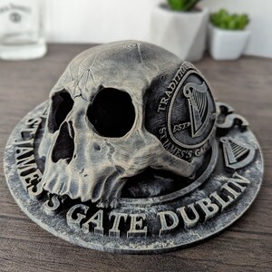 Może przedstawiać: Szary przedmiot dekoracyjny w kształcie czaszki z okrągłą podstawą. Na podstawie znajduje się napis "ST JAMES'S GATE DUBLIN" oraz logo z harfą. Czaszka ma szczeg&oacute;łowe detale i spoczywa na podstawie.