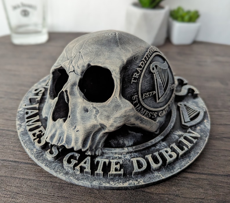 Może przedstawiać: Szary przedmiot dekoracyjny w kształcie czaszki z okrągłą podstawą. Na podstawie znajduje się napis "ST JAMES'S GATE DUBLIN" oraz logo z harfą. Czaszka ma szczeg&oacute;łowe detale i spoczywa na podstawie.