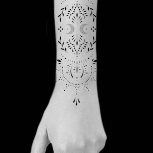 Puede incluir: Diseño de tatuaje en blanco y negro en el antebrazo. El tatuaje presenta un patrón simétrico con elementos florales y de media luna. El diseño incluye puntos, líneas y formas de hojas, creando una estética intrincada y detallada.