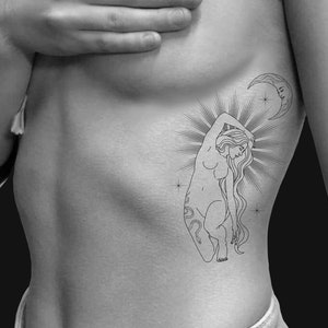 Puede incluir: Tatuaje en blanco y negro de una mujer con el pelo largo y suelto, arrodillada con el brazo levantado. También se representa una luna creciente con una cara y un sol radiante. El tatuaje está en el costado del torso de una persona.