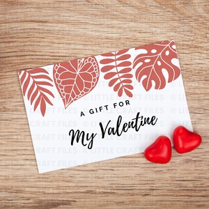 May include: A rectangular white card featuring a terracotta-colored botanical leaf pattern along the top edge and the black text A GIFT FOR My Valentine. Two small, shiny red heart-shaped candies are positioned near the bottom right corner.