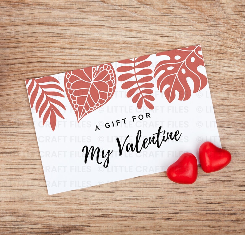 May include: A rectangular white card featuring a terracotta-colored botanical leaf pattern along the top edge and the black text A GIFT FOR My Valentine. Two small, shiny red heart-shaped candies are positioned near the bottom right corner.