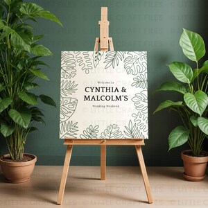 May include: A square white sign on a wooden easel featuring a black line art border of tropical leaves. The text reads Welcome to CYNTHIA & MALCOLM'S Wedding Weekend in black lettering. The sign is positioned between two potted green plants.