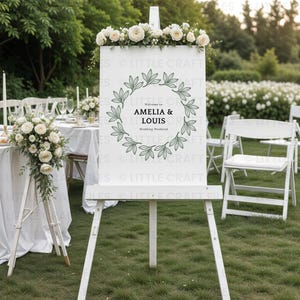 May include: A white rectangular wedding welcome sign displayed on a wooden easel, featuring a dark green botanical leaf wreath graphic surrounding the text Welcome to AMELIA & LOUIS Wedding Weekend, topped with a floral arrangement of white roses.