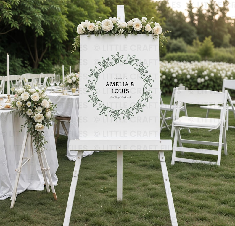 May include: A white rectangular wedding welcome sign displayed on a wooden easel, featuring a dark green botanical leaf wreath graphic surrounding the text Welcome to AMELIA & LOUIS Wedding Weekend, topped with a floral arrangement of white roses.