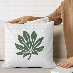 May include: A square white throw pillow featuring a centered, large, dark green botanical leaf graphic with distinct pointed lobes and a central vein structure. The pillow has a smooth fabric texture and a clean, simple design.
