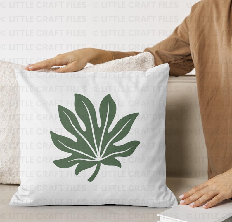 May include: A square white throw pillow featuring a centered, large, dark green botanical leaf graphic with distinct pointed lobes and a central vein structure. The pillow has a smooth fabric texture and a clean, simple design.