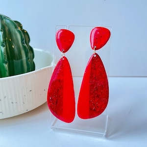 May include: A pair of red dangle earrings with a teardrop shape. The earrings have a metallic gold fleck embedded in the red resin.