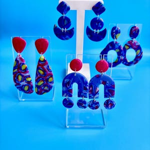 Op de afbeelding: Een verzameling kleurrijke polymeerklei oorbellen. De oorbellen hebben verschillende vormen, waaronder druppels, bogen en geometrische ontwerpen, met levendige patronen in rood, geel en blauw. Sommige oorbellen hebben een effen rode accent.