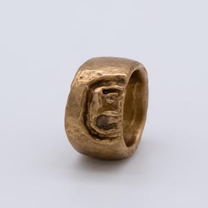 Op de afbeelding: Een bronzen ring met een getextureerd oppervlak. De ring heeft een dikke band en een gebeeldhouwd ontwerp aan de voorkant, mogelijk een gezicht of figuur. De ring staat tegen een witte achtergrond.