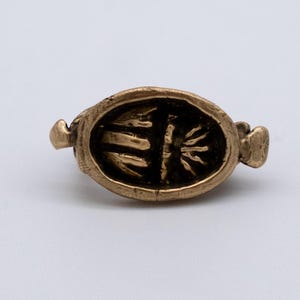 May include: A small, antique-style ring with an oval face. The ring is made of a gold-colored metal and features an engraved design within the oval. The design appears to be a stylized depiction of a plant or symbol. The ring has small, rounded extensions on either side.