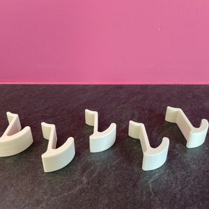 Puede incluir: Cinco ganchos de pl&aacute;stico blanco, dise&ntilde;ados para colgar objetos, est&aacute;n dispuestos en una fila sobre una superficie gris oscuro.