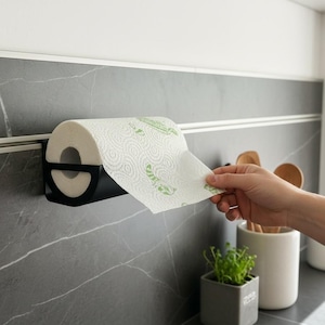 Könnte beinhalten: Ein schwarzer Papierhandtuchhalter an einer grau gefliesten Wand. Eine Rolle weißer Papierhandtücher mit grünem Blättermuster befindet sich im Halter. Eine Hand zieht ein Blatt Papierhandtuch von der Rolle.