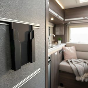 Könnte beinhalten: Ein schwarzer, an der Wand befestigter Organizer mit zwei Fächern, befestigt an einer strukturierten grauen Wand mit silberner Verkleidung. Im Hintergrund ist das Innere eines Wohnmobils mit Küche, Sitzgelegenheiten und einem Fenster zu sehen.