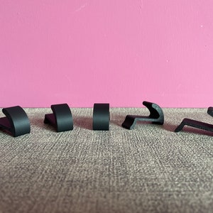 Puede incluir: Seis ganchos de pl&aacute;stico negro, algunos con forma curva, dispuestos en fila sobre una superficie gris texturizada.