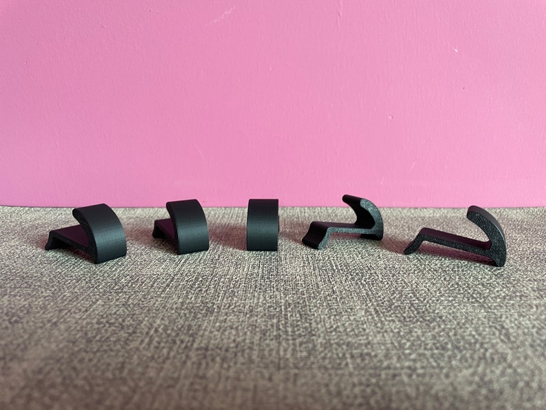 Puede incluir: Seis ganchos de pl&aacute;stico negro, algunos con forma curva, dispuestos en fila sobre una superficie gris texturizada.